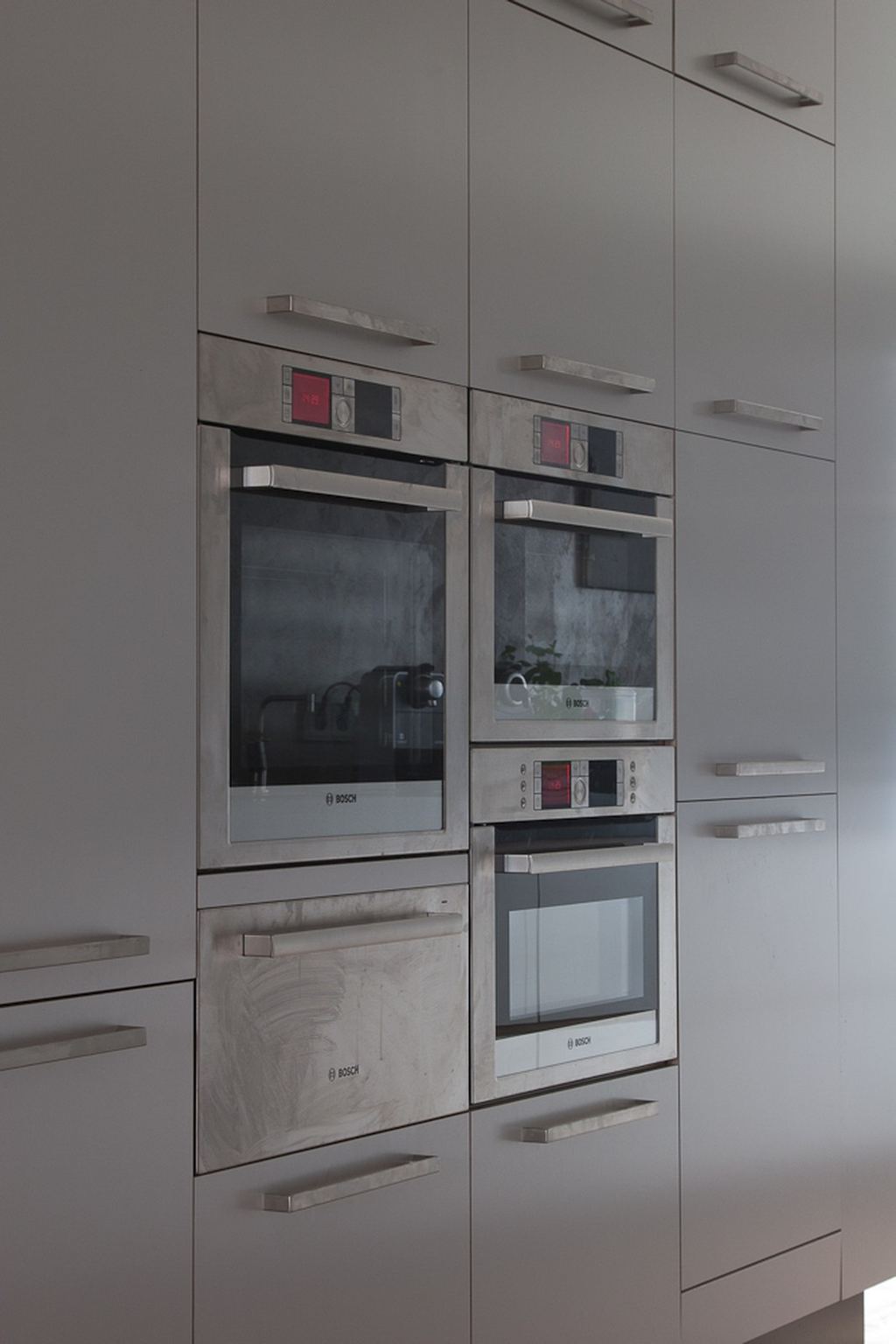 Schuin gefotografeerde kastenwand met ingebouwde ovens en lange, smalle metalen handgrepen.