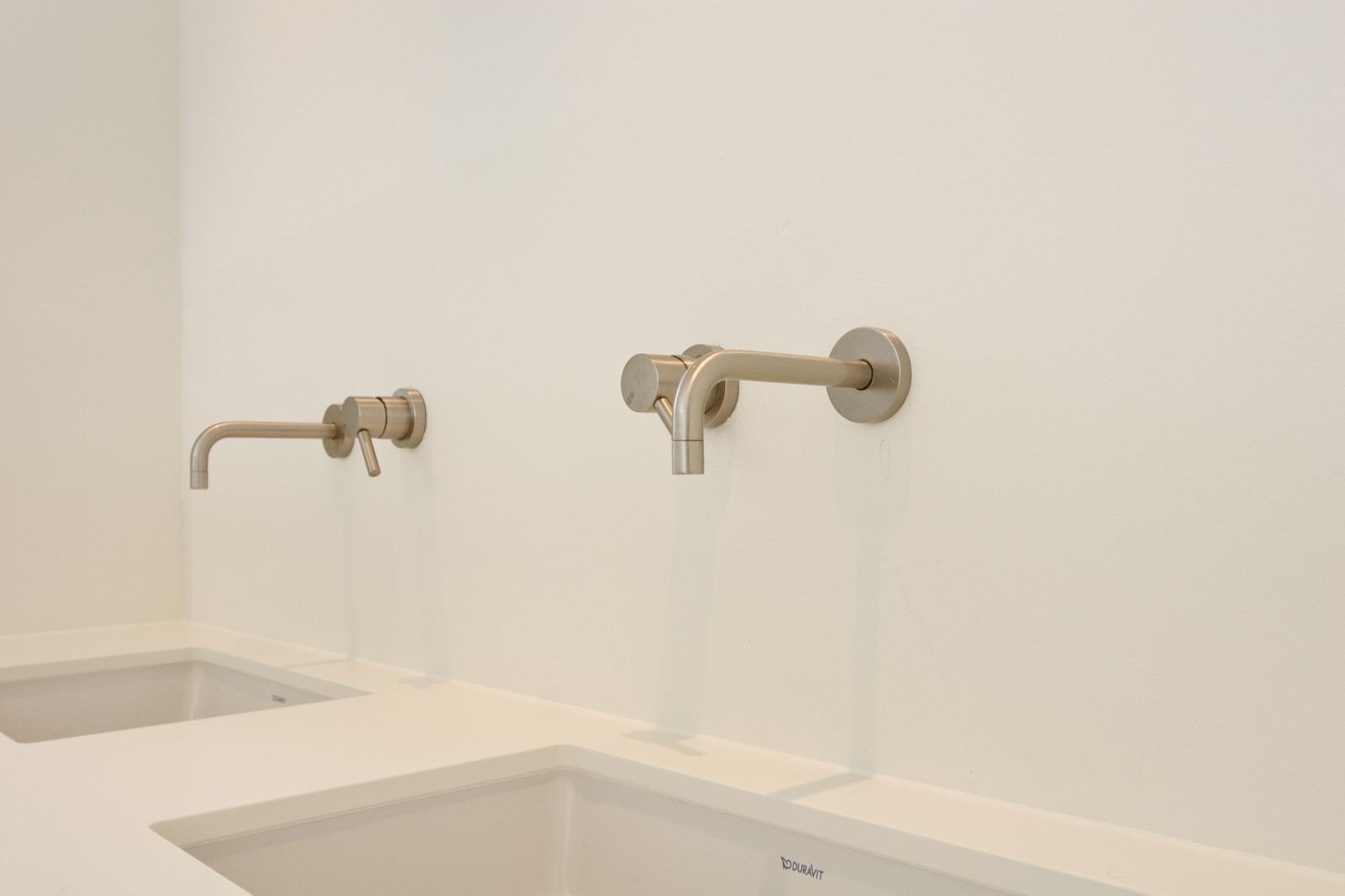 Twee wandkranen boven het wit spuitwerk wastafelmeubel.