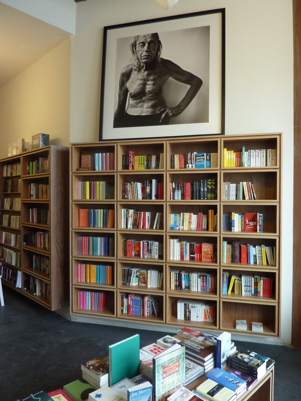 Vierkant boekenrek van licht hout, onder een groot ingelijst portret.